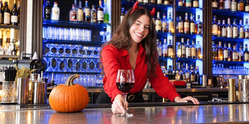 Bartender in a red shirt and devil horns serving red wine beside a pumpkin on the bar.