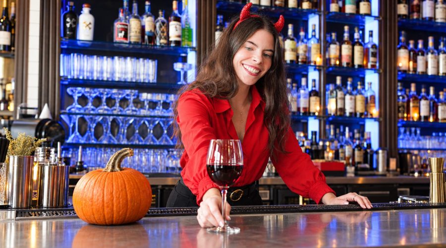 Bartender in a red shirt and devil horns serving red wine beside a pumpkin on the bar.