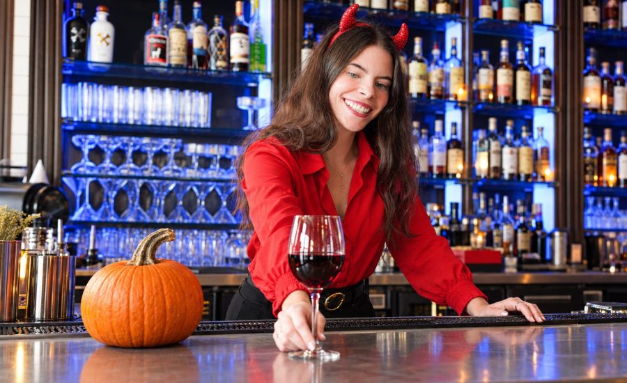 Bartender in a red shirt and devil horns serving red wine beside a pumpkin on the bar.
